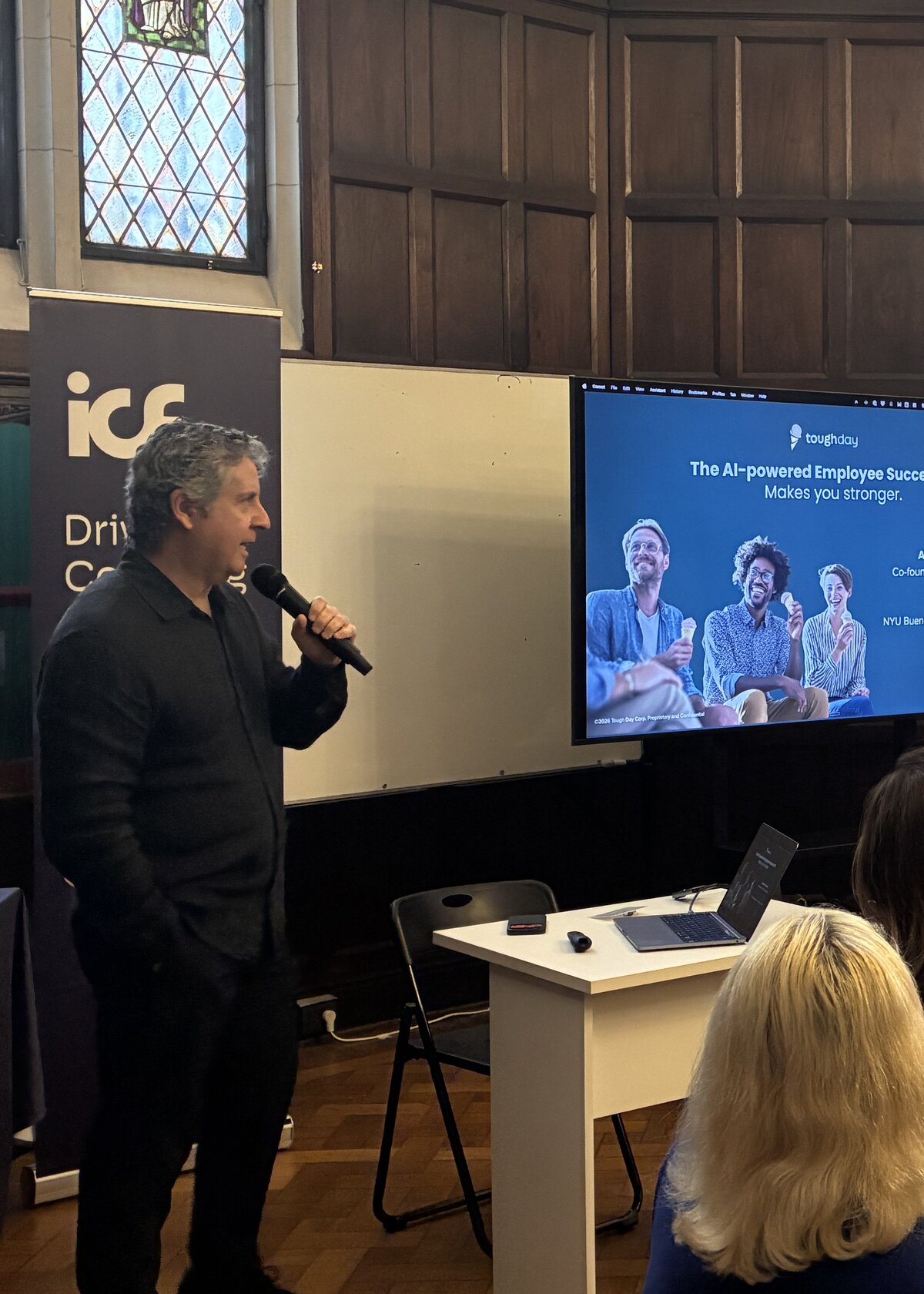 Alberto holding a microphone at NYU Buenos Aires, next to a screen showing the Tough Day 'AI-powered Employee Success OS' slide